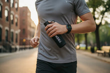 Charger l'image dans la galerie, Homme courant en ville tenant une gourde noire TREK, portant un t-shirt gris et une montre connectée